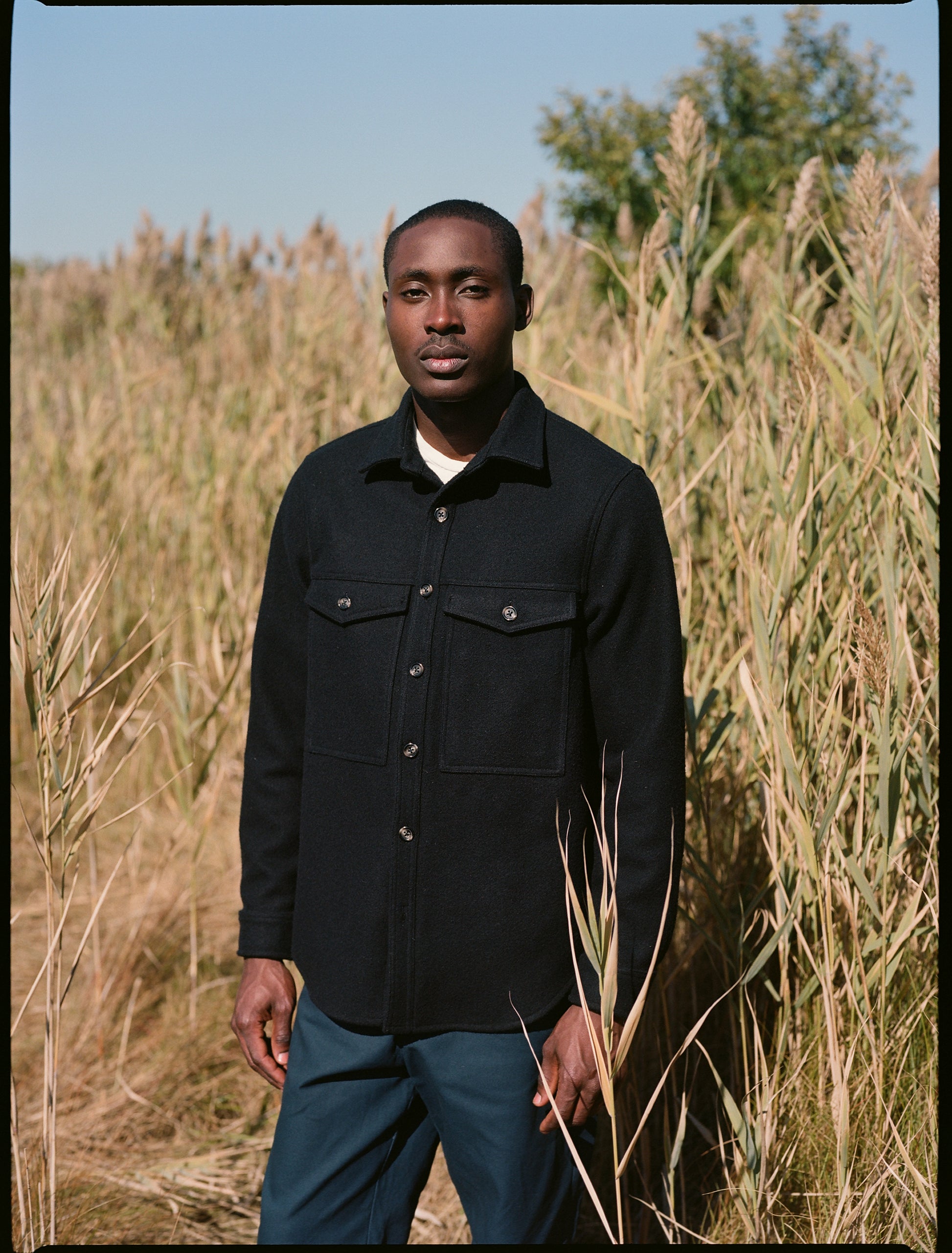 Model standing up in a field, wearing black melton wool double pocket shirt jacket and navy danver pants 