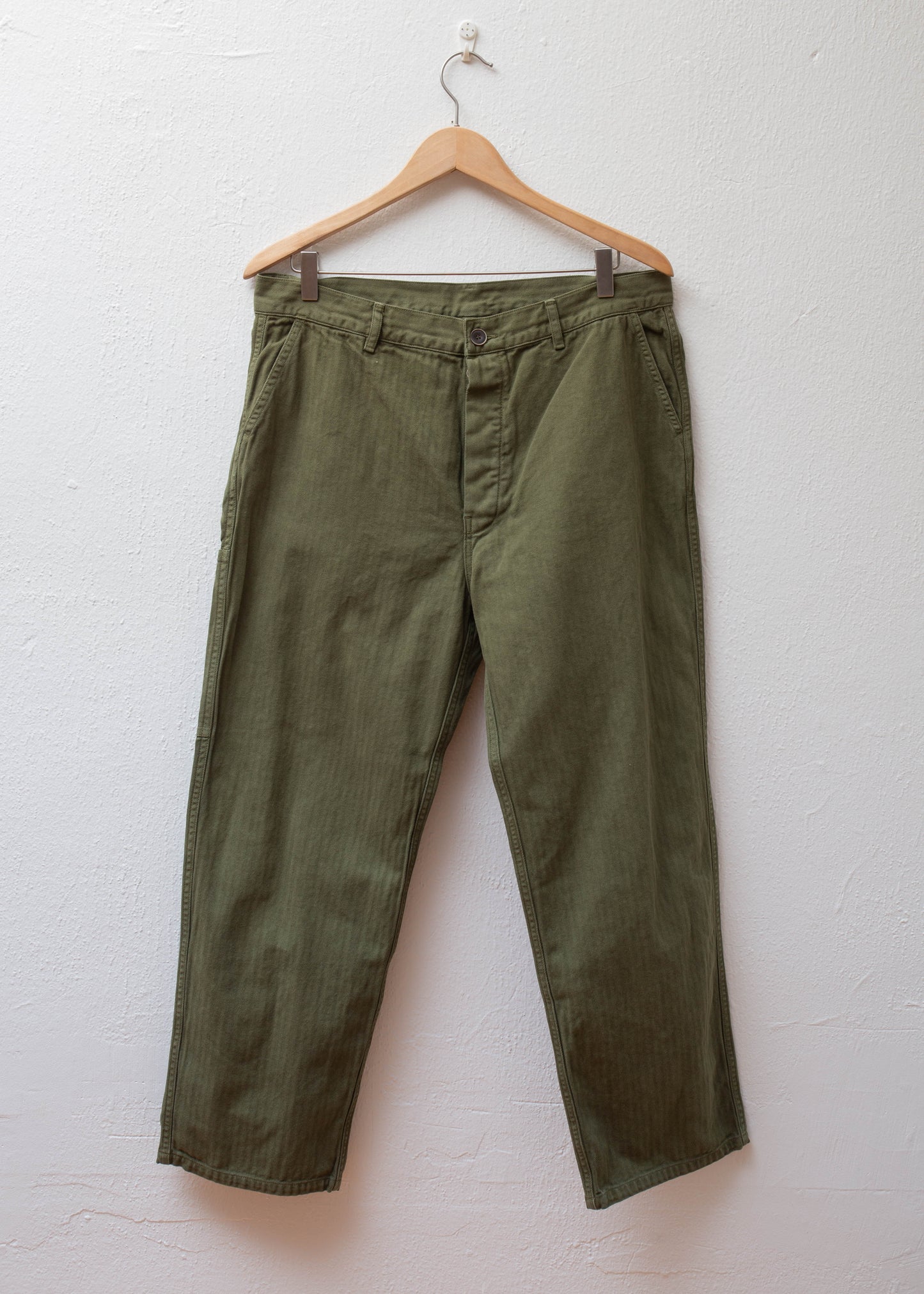 Herringbone Over Dye Pants color Olive hanging on white wall 